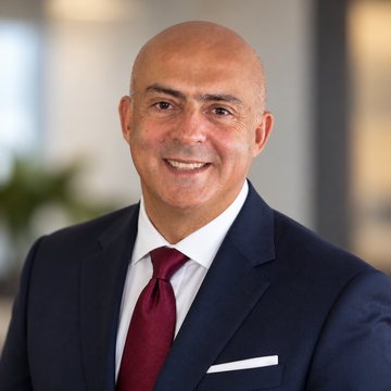 Jose Salloum, Infinite Banking practitioner, in a navy suit and a burgundy tie with a pocket square, a plant behind him