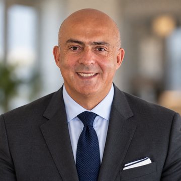 Jose Salloum, Infinite Banking practitioner, in a charcoal suit and a navy tie against a plain grey wall